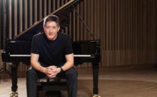 A pianist sits on a piano bench in a modern concert hall, hands clasped, with a grand piano behind h