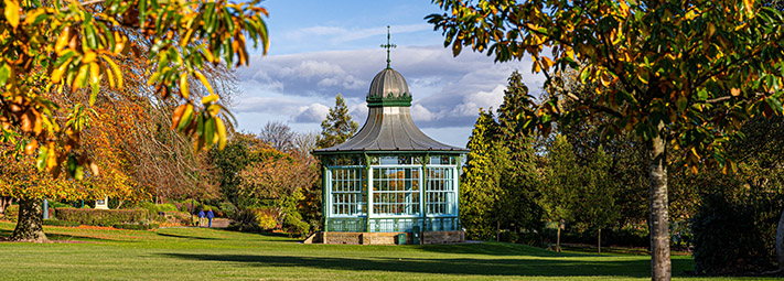 A bandstand is bathed in sunlight under a blue sky, surrounded by leafy greenery in a park