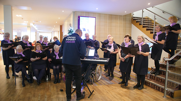 Photo of OutSingCancer choir rehearsing with keyboardist wearing jumper saying "united against cancer" on the back of it.
