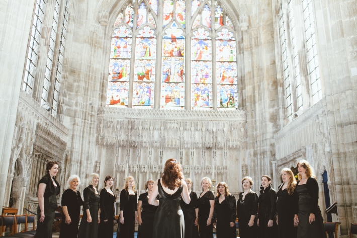 La Nova Singers female choir performing in Christchurch Priory. The singers wear elegant black evening gowns and sing in front of a large stained glass window