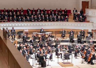 members of the choir and orchestra on stage, sitting down, ready to being the concert