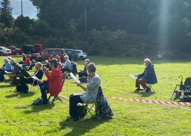 seated choir have a socially distanced rehearsal in green space outdoors