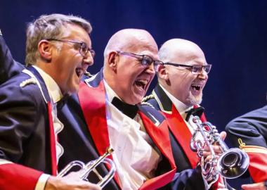 Three male members of Derwent Brass on stage, laughing and smiling whilst performing Brassed Off at Derby Theatre in red and navy suits.