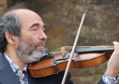 Man wearing grey suit, holding violin, pictured outside