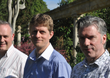 3 smiling men wearing open necked shirts, pictured outside
