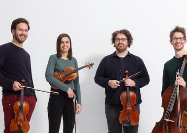 Four young people dressed in black standing in front of a white wall, holding string instruments 