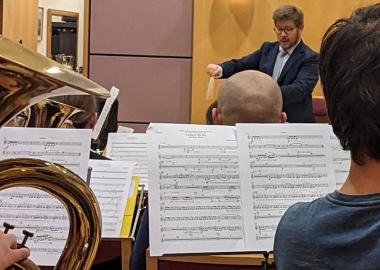 close-up of music sheets with students playing, with music director conducting in the background