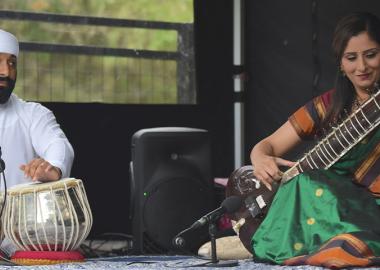 Concert performance featuring sitar player Roopa Panesar and tabla player Rajvir Bhachu. Roopa is seated on the right in a green and maroon sari, playing the sitar, while Rajvir, dressed in white with a white turban, plays the tabla on the left. They are performing on a stage with microphones and a speaker in the background. 