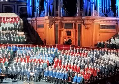 Photo of Welsh Male choir performing at the Royal Albert Hall. The singers are wearing red, black, and blue attire. There is an organ in the background and a pianist and conductor in the foreground 