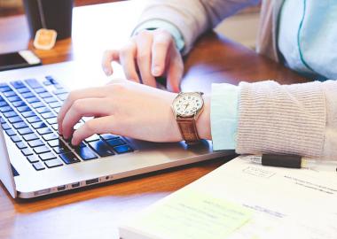 Hands typing on a MacBook laptop, wearing a watch on one hand and a notebook and pen lays underneath that same hand.