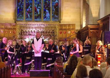 musicians of NULCO sit with their instruments in a church while their MD addresses the audience