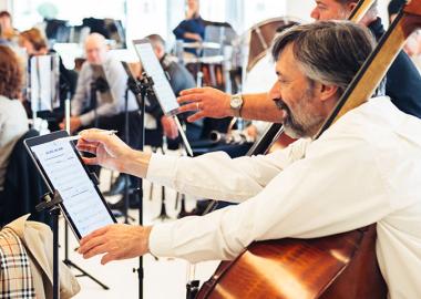 A man with a bass violin resting on his shoulder as he annotates sheet music on an iPad.